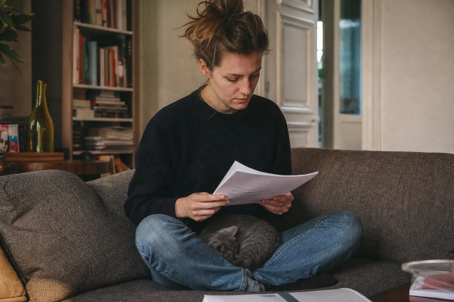 Une jeune femme lit un contrat d'assurance sur son canapé, un chaton gris endormi contre elle