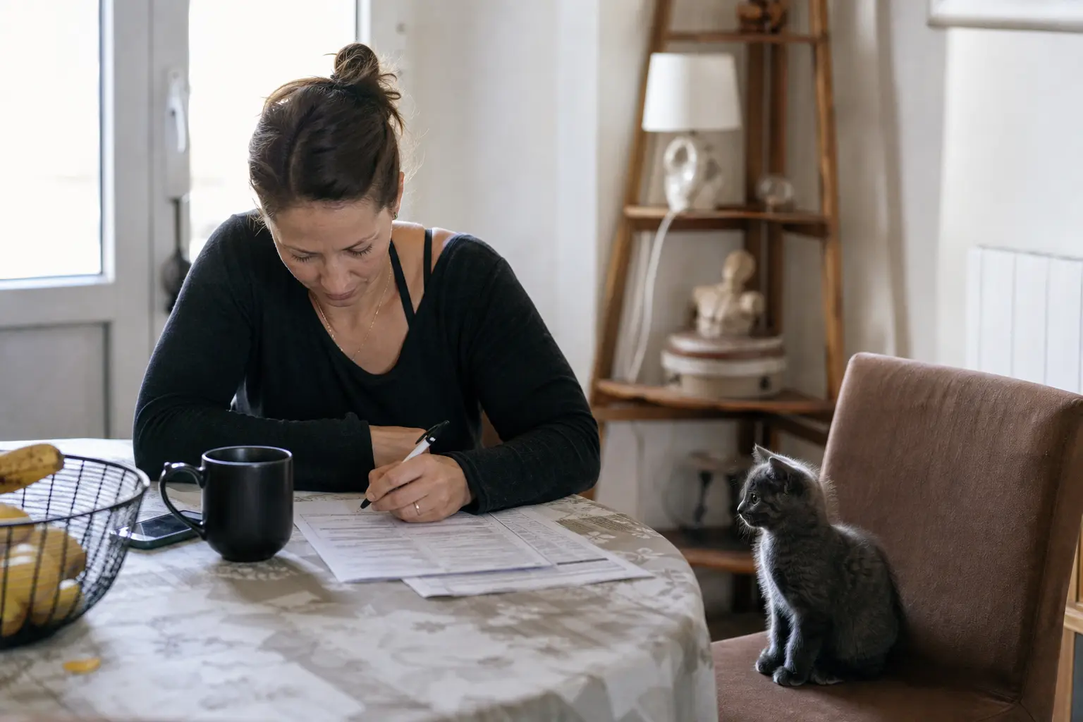 Une femme remplit un formulaire papier à sa table, un chaton assis sur la chaise voisine