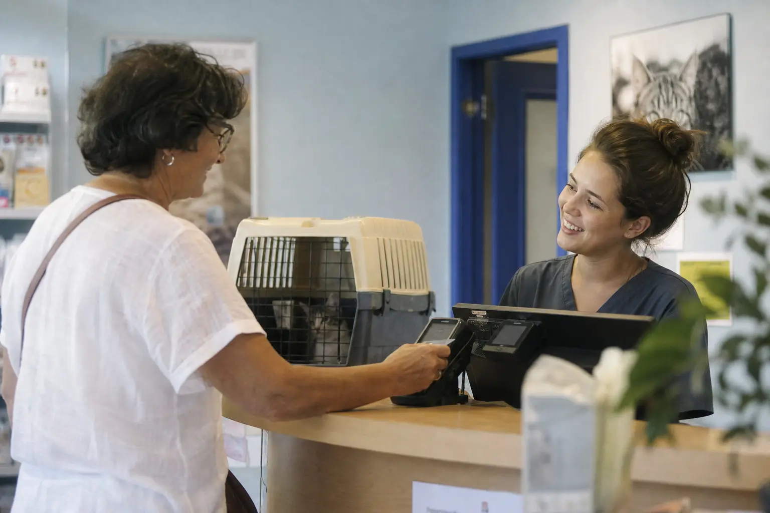 Une femme règle à l'accueil d'une clinique vétérinaire, caisse de transport posée sur le comptoir